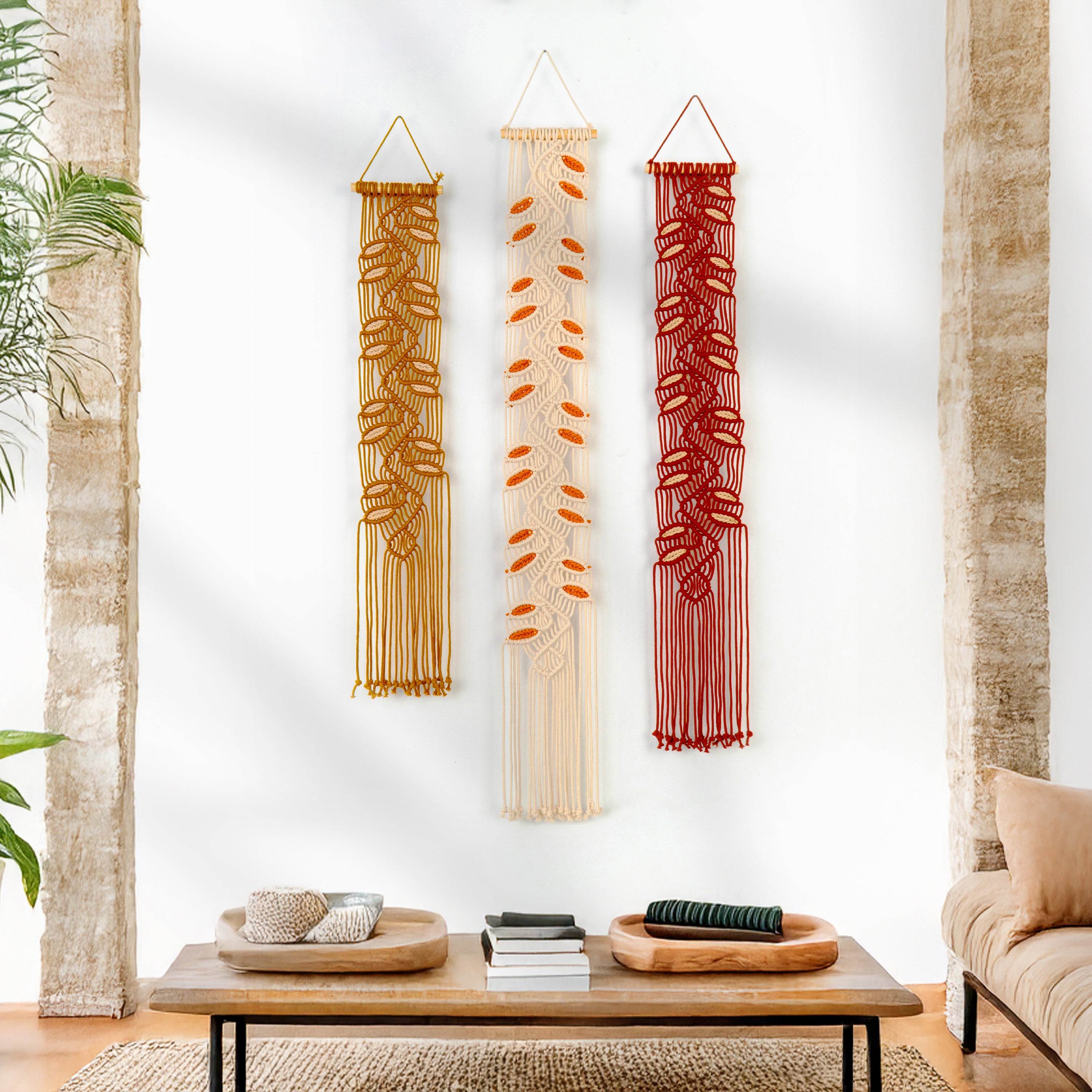 Three macrame wall hangings in gold, beige, and red hanging on a white wall above a wooden coffee table.