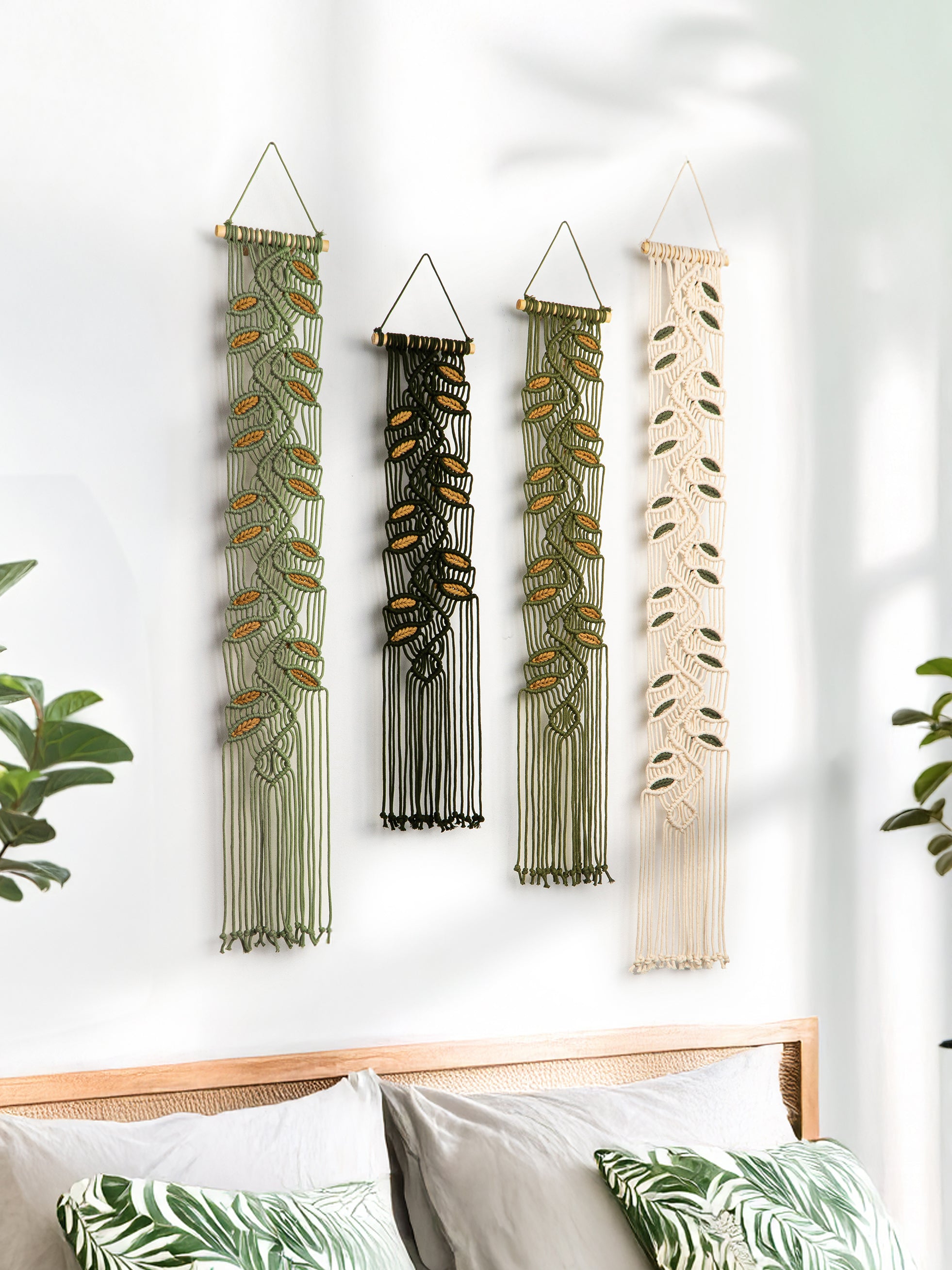 Four macrame wall hangings with leaf patterns on a white wall above a bed.