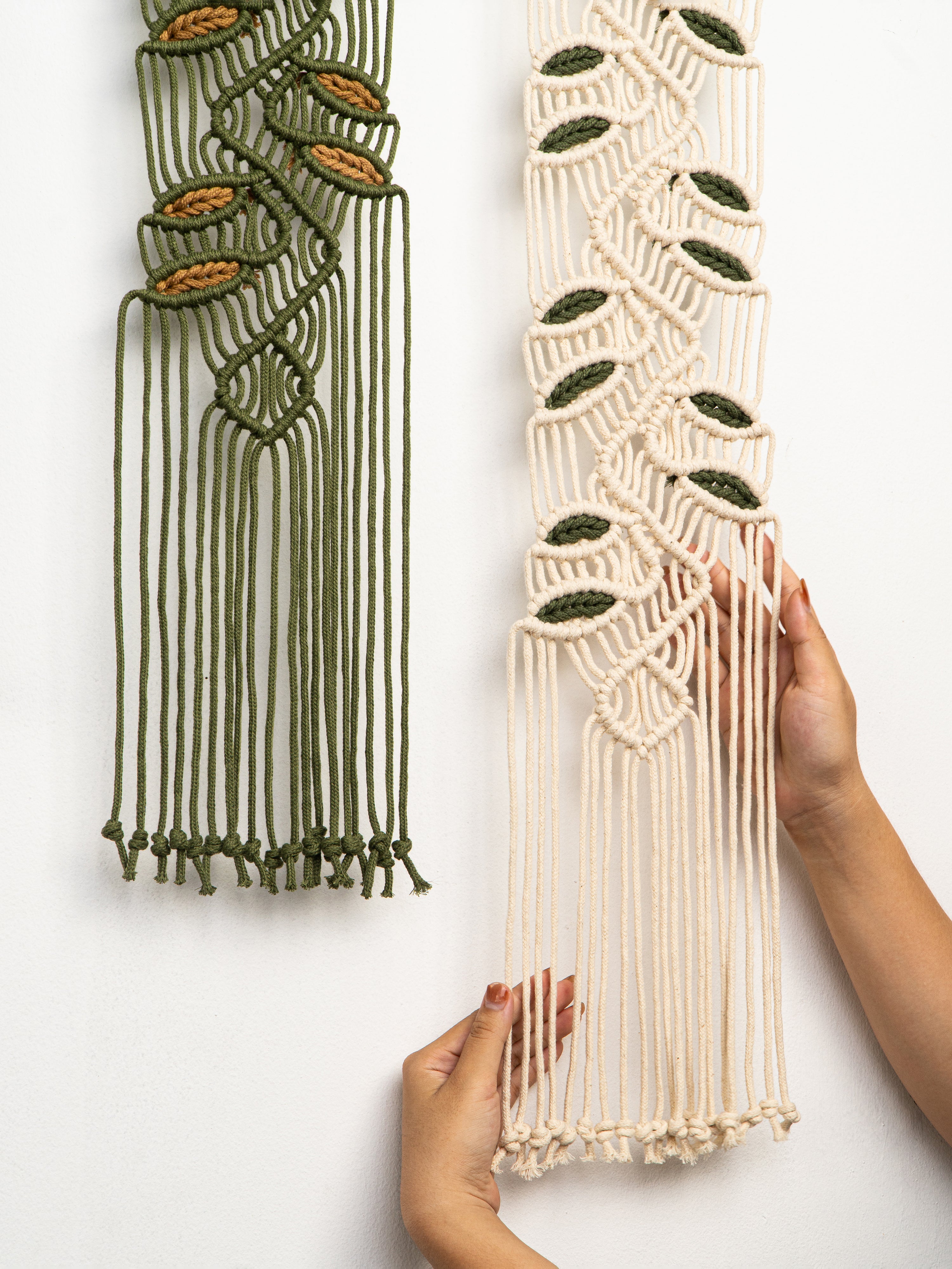 Two macrame wall hangings, one green and one beige, held by hands against a white background.