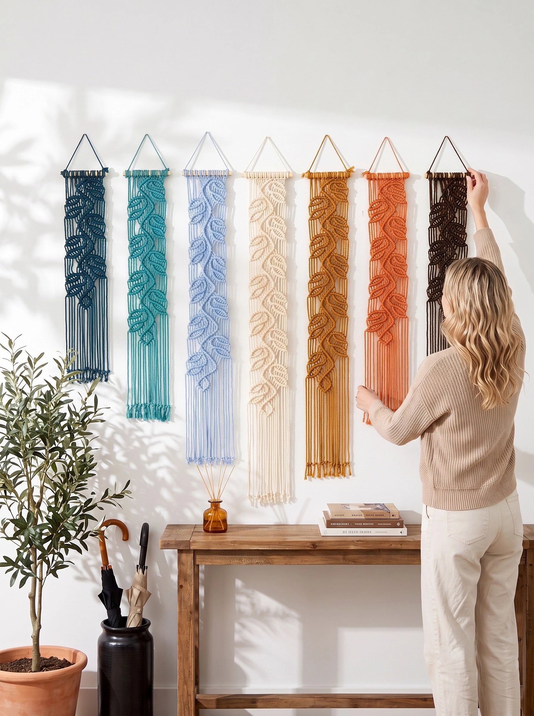 A woman hangs a large woven macrame wall hanging on a white wall in a bright room. Six additional woven wall hangings in teal, light blue, cream, tan, rust, and dark brown are displayed on the wall above a wooden console table with a potted plant and small decor items.