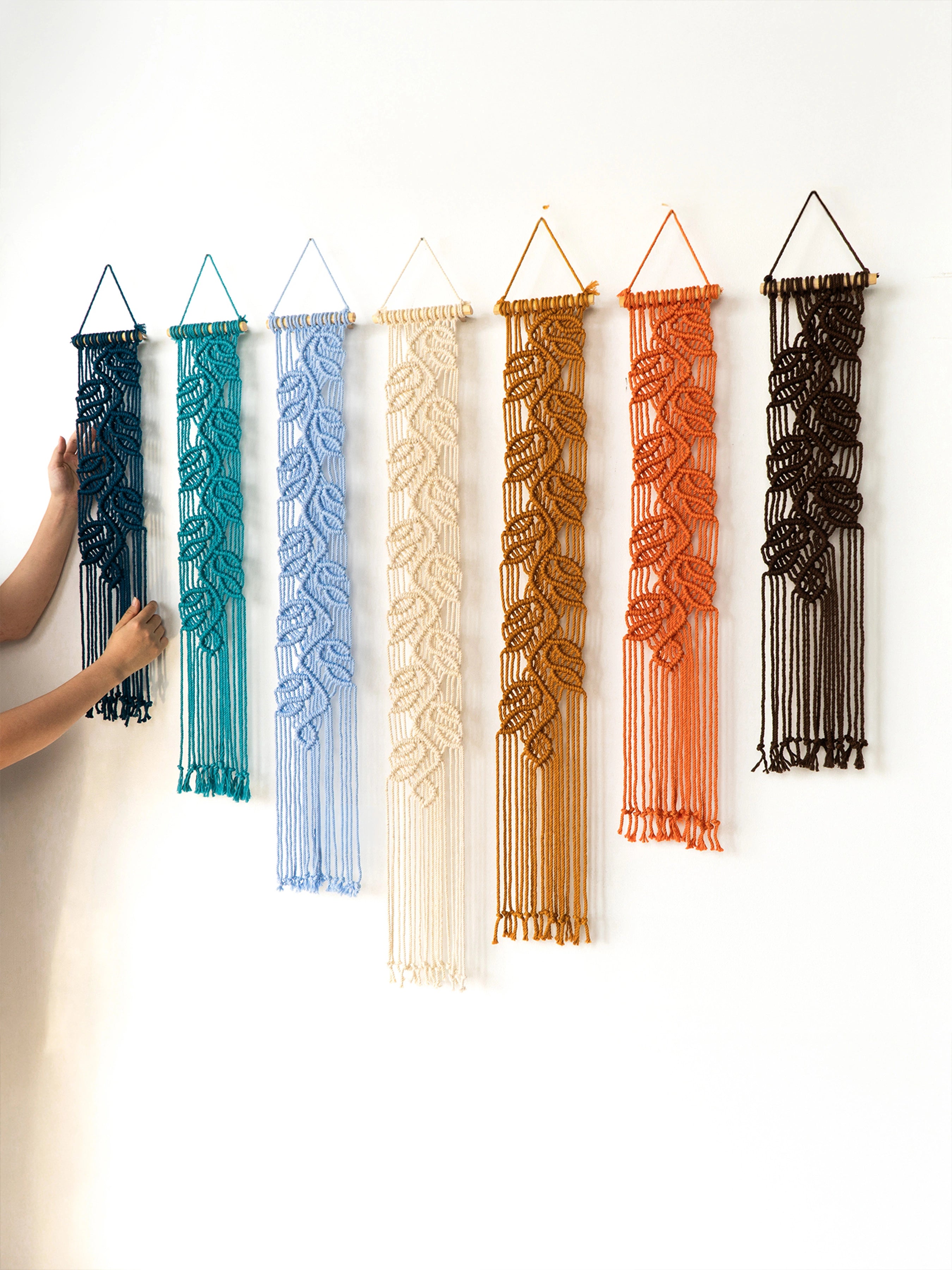 Six woven macrame wall hangings in different colors are displayed on a plain white wall. A person's arm and hand are visible on the left, adjusting the first hanging.