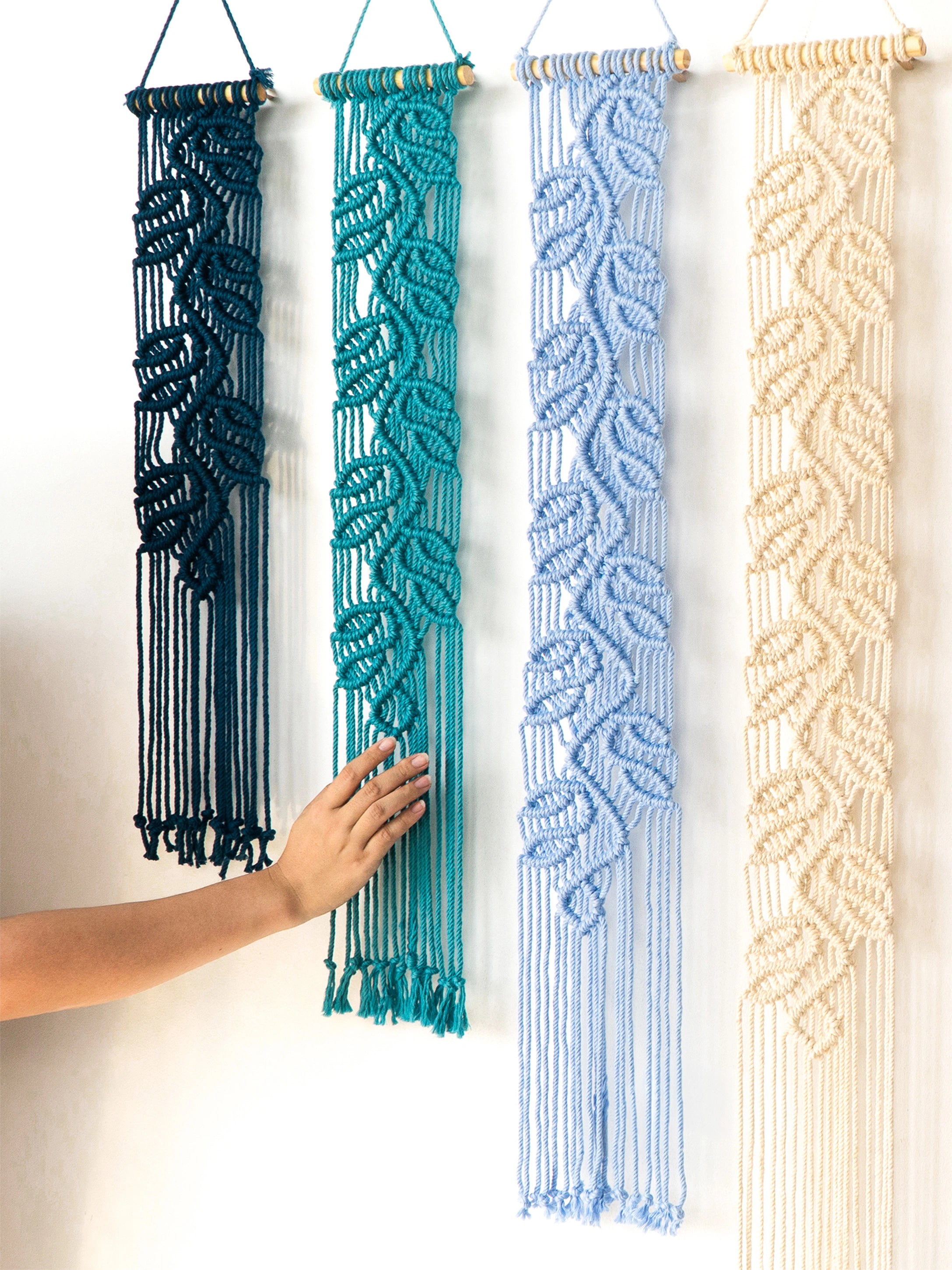 Four macrame wall hangings in dark teal, turquoise, light blue, and cream are displayed against a white wall, with a hand touching the turquoise one.