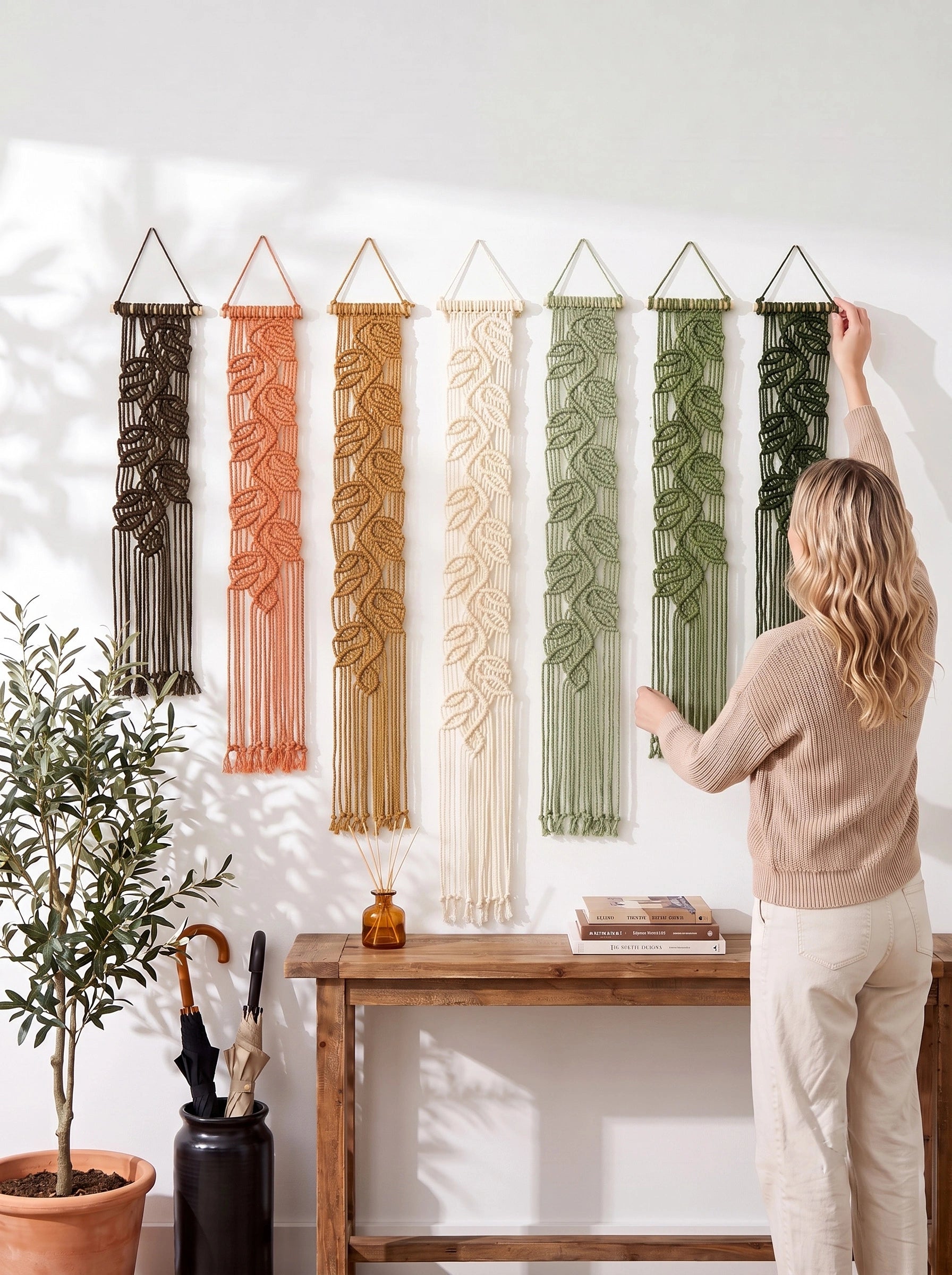 A woman hangs a green macrame wall hanging on a white wall next to a wooden console table. Five other macrame wall hangings in brown, orange, cream, sage green, and dark green are displayed on the wall above the table, with a potted plant and decorative items on the floor.