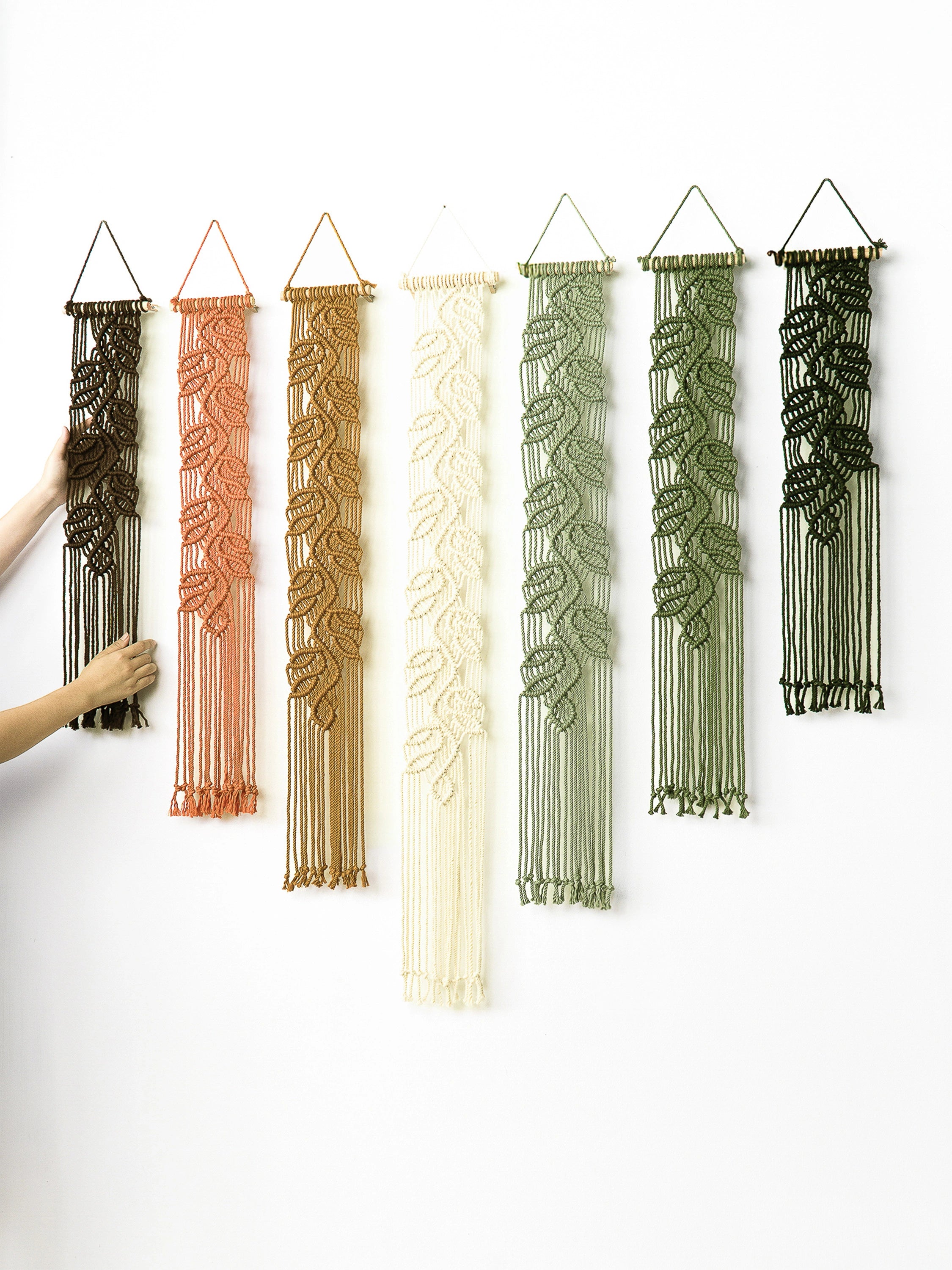Six woven macrame wall hangings with leaf patterns in black, terracotta, tan, cream, sage green, and olive green, displayed on a white wall. A person's arm and hand are visible on the left adjusting the first hanging.