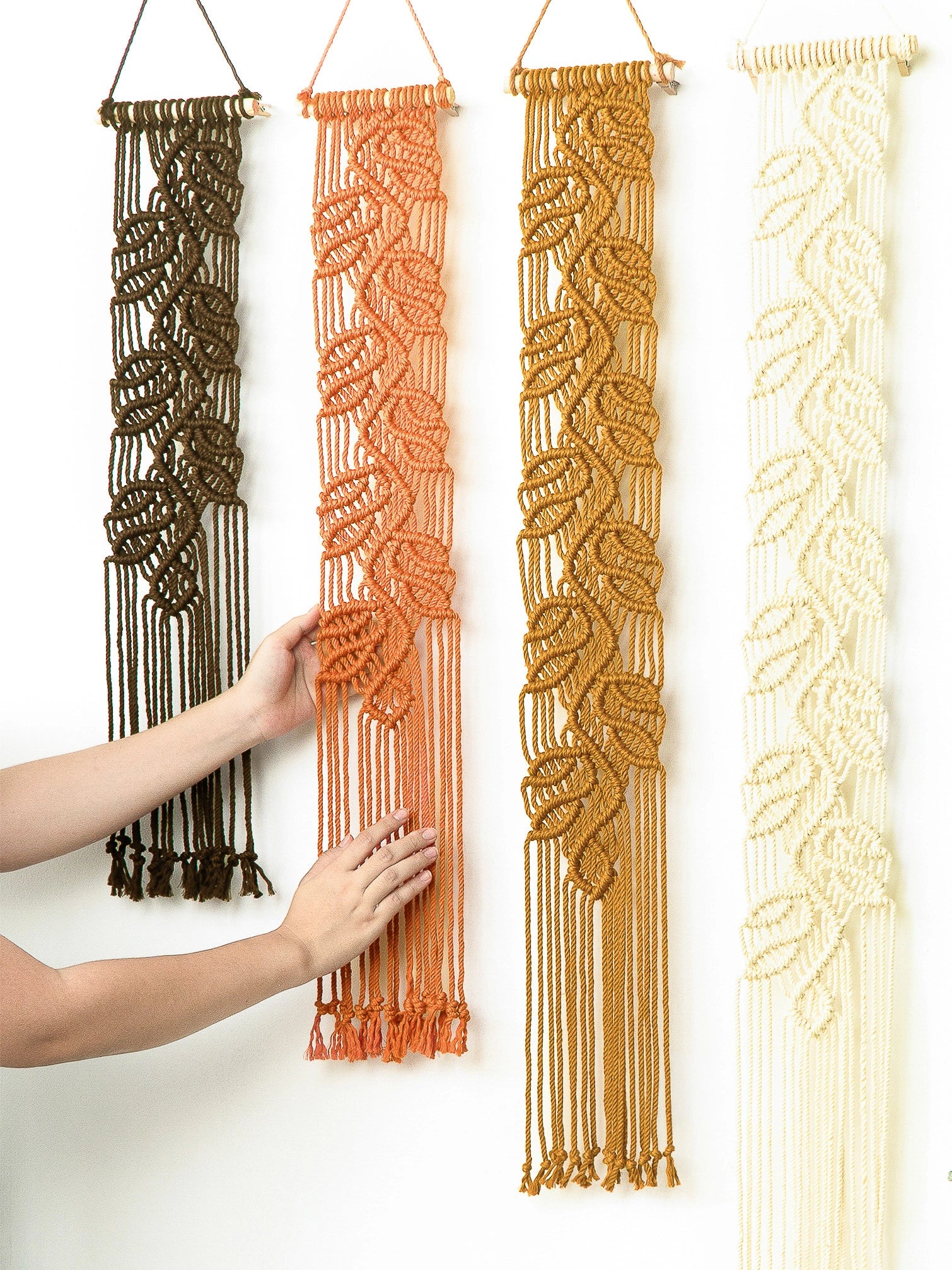 Four vertical macrame wall hangings in dark brown, terracotta orange, mustard yellow, and cream are displayed against a white wall. A person's hands are visible holding the center orange hanging to show its texture and scale.