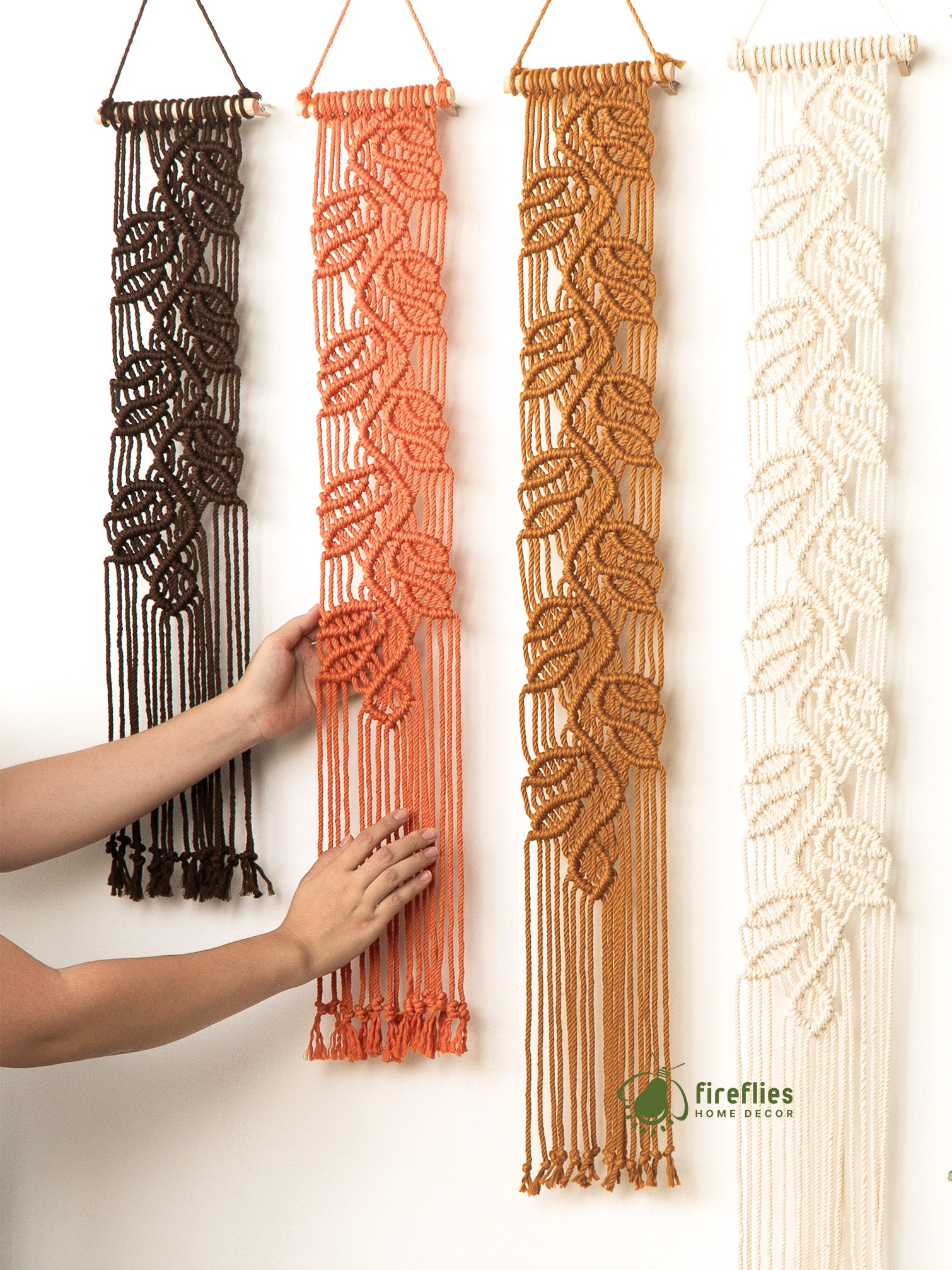 Four macrame wall hangings in different colors being held by a person against a white background.