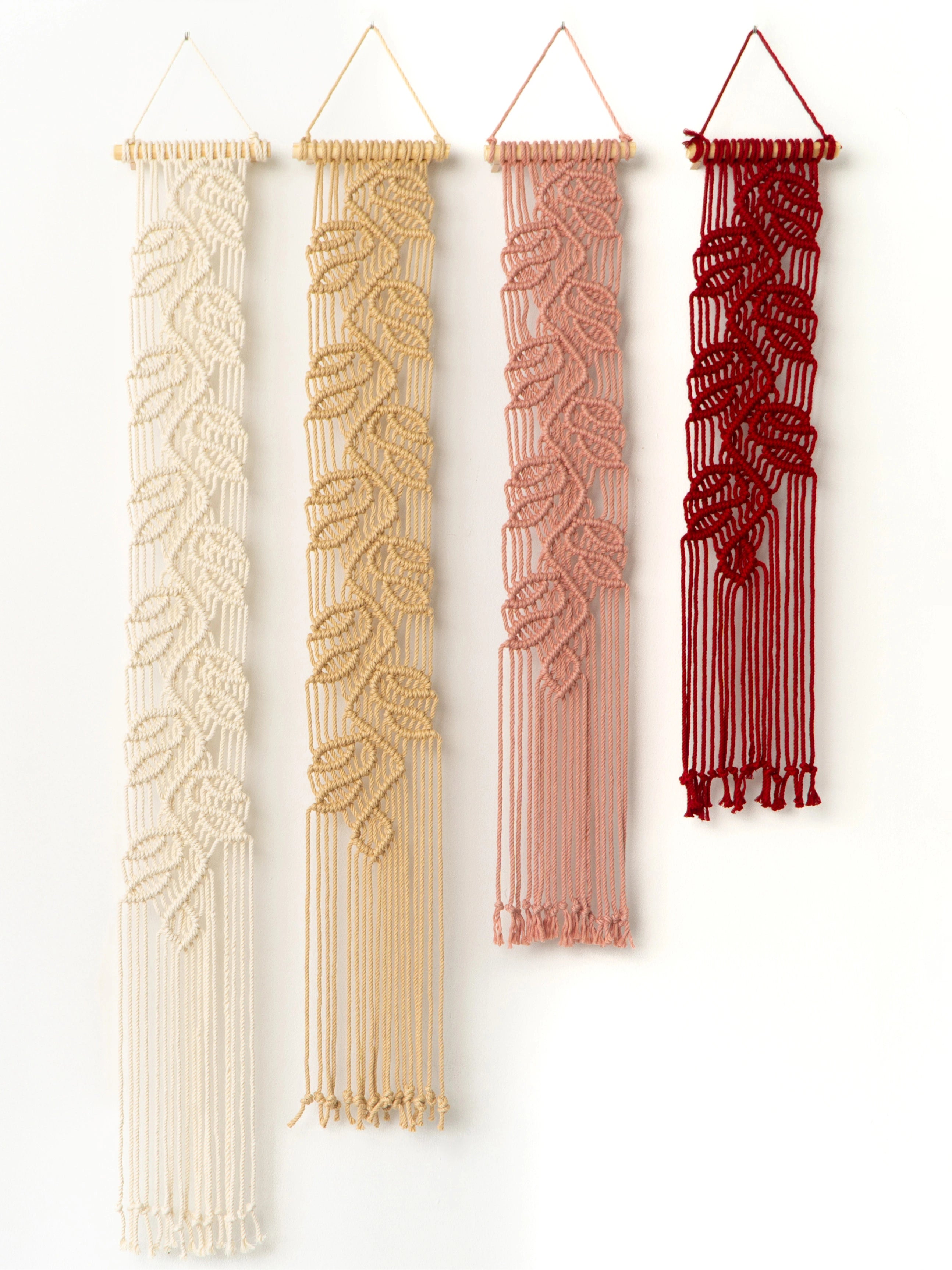 Four bohemian-style macrame wall hangings with leaf patterns in cream, tan, dusty pink, and deep red, displayed vertically against a plain white wall.