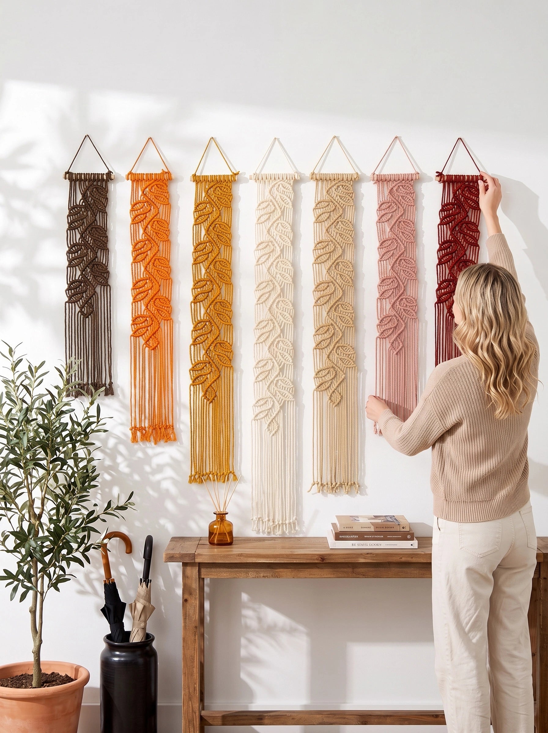 A woman hangs a red macrame wall hanging on a white wall next to five other macrame wall hangings in brown, orange, cream, beige, and pink. A wooden console table, a potted plant, and a vase with umbrellas are below.