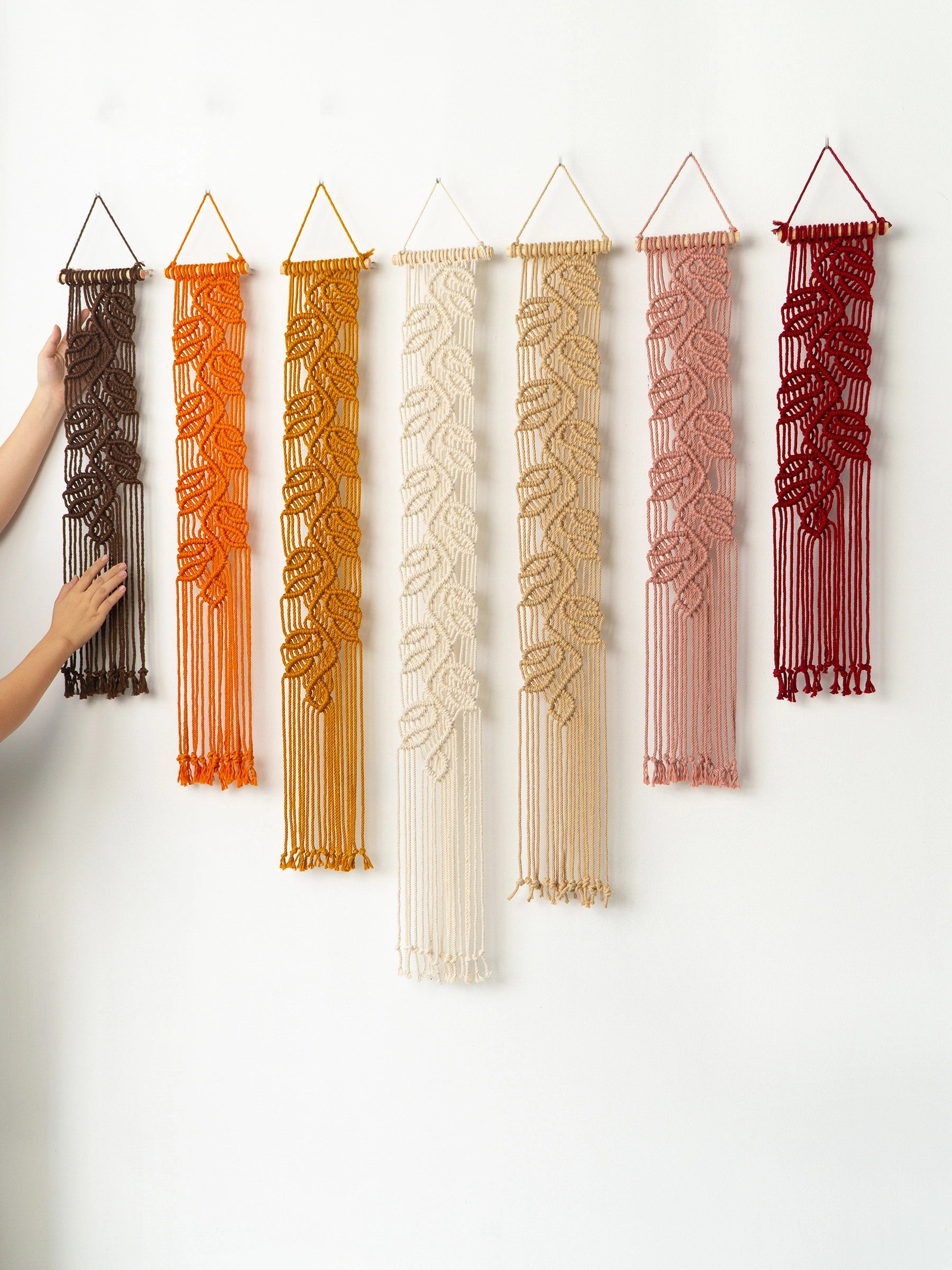 Six woven macrame wall hangings in brown, orange, mustard, cream, blush pink, and burgundy are displayed on a white wall, with a person's hands touching the brown one on the left.