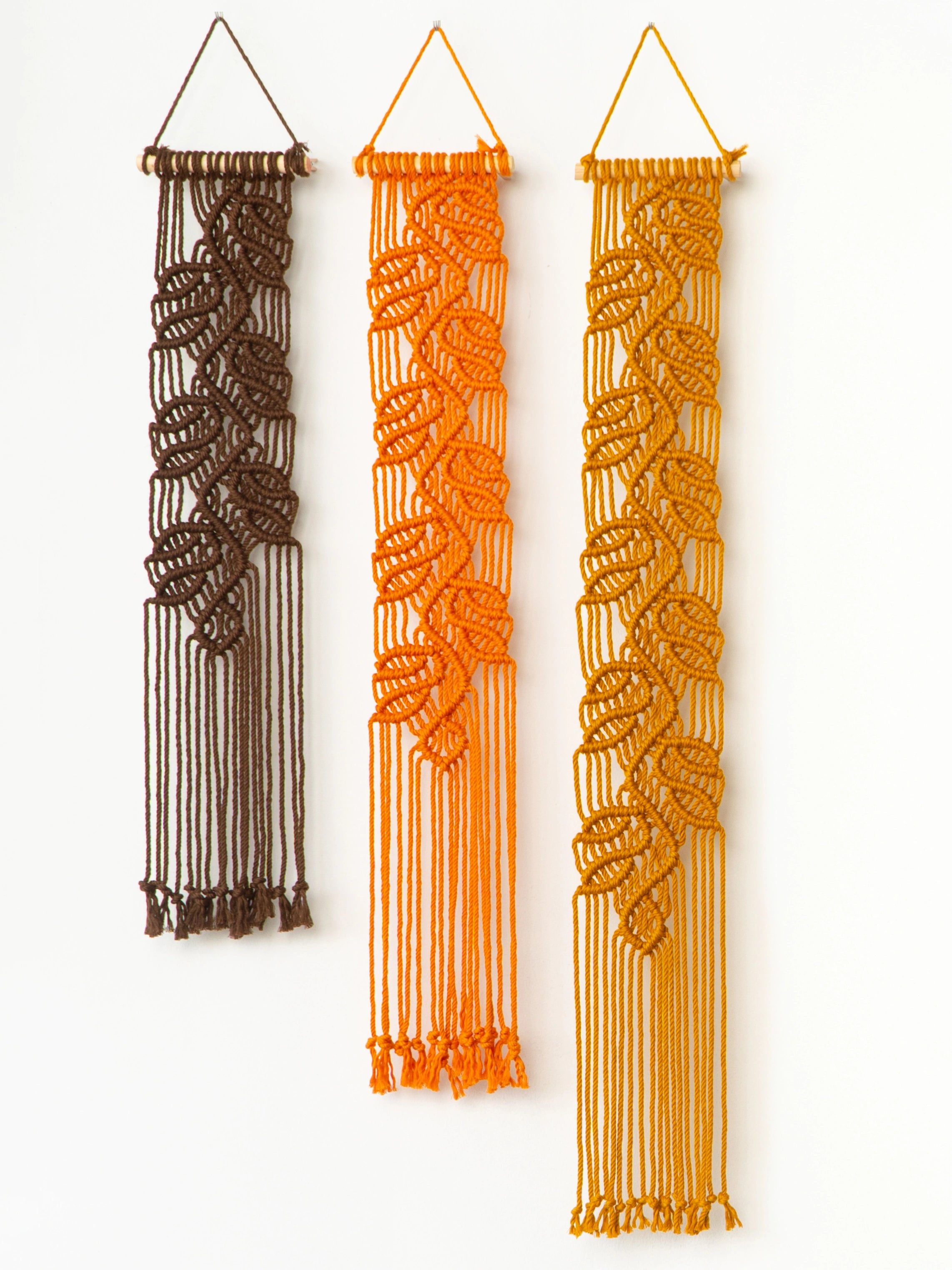 Three vertical macrame wall hangings with leaf-like woven patterns, shown hanging against a plain white wall. The left one is dark brown, the middle one is orange, and the right one is golden yellow.