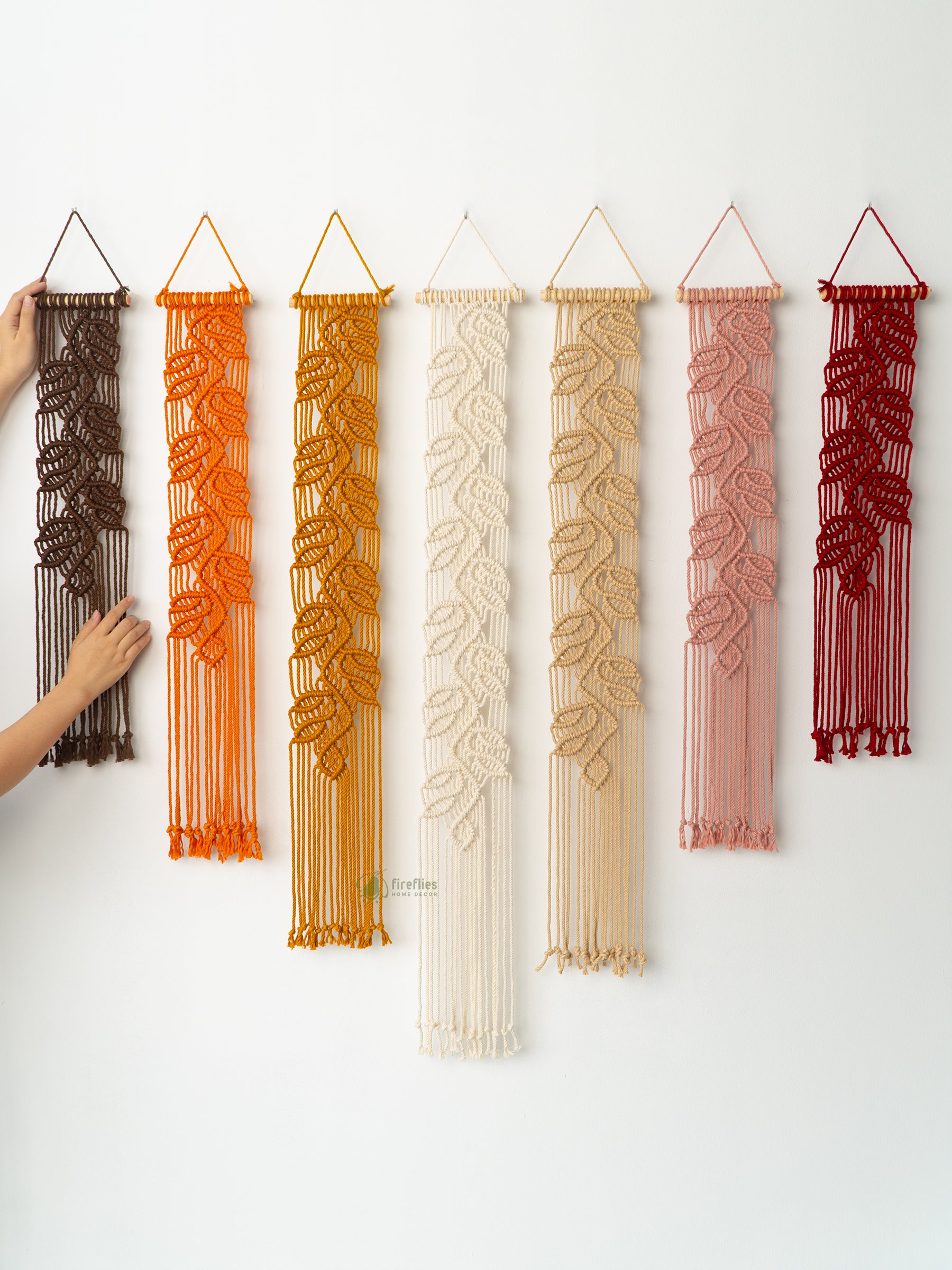 Artisan fiber art close-up: four macrame leaf hangings suspended from wooden dowels against a white wall.