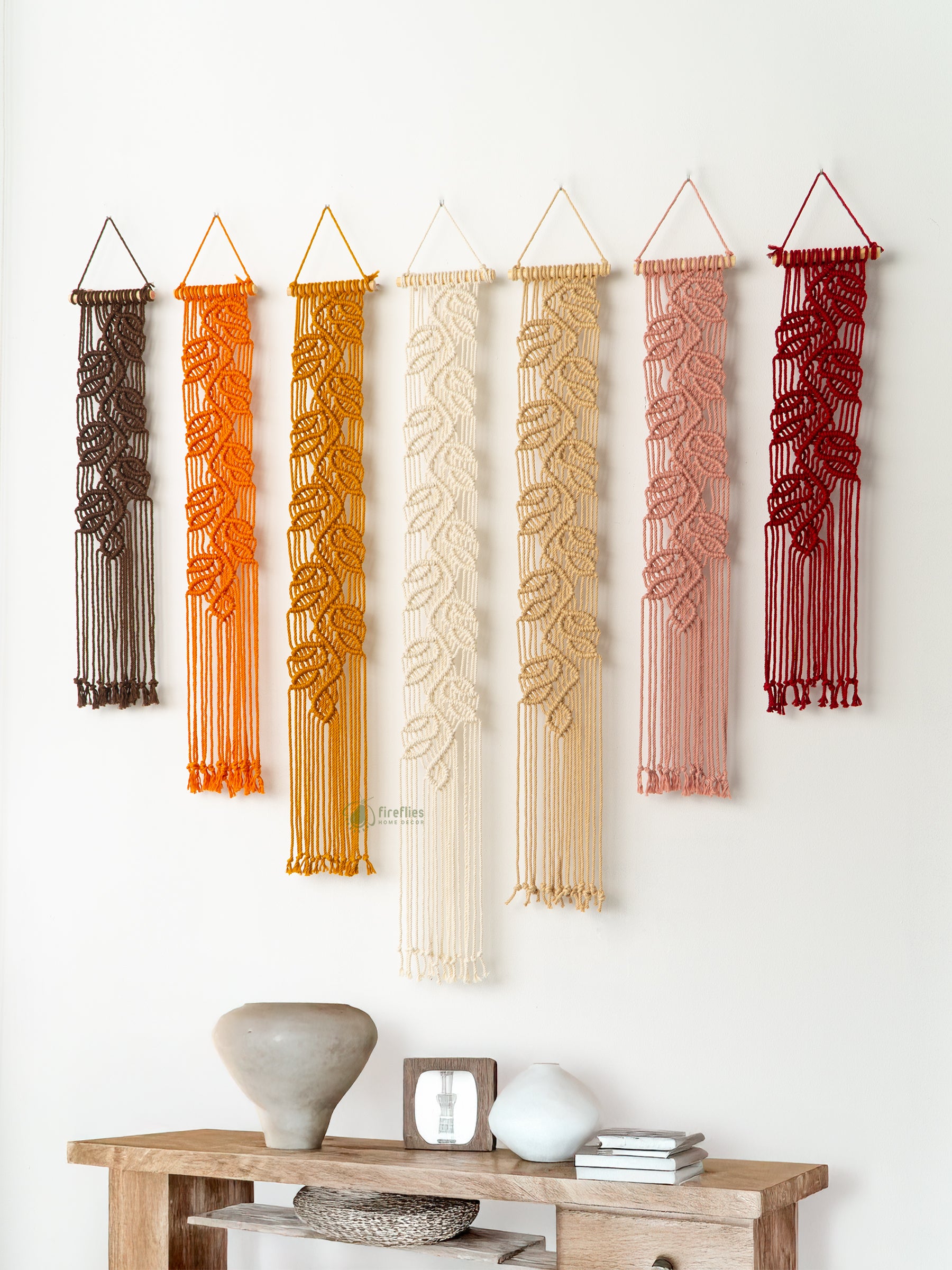Set of six macrame wall hangings in various colors hanging on a white wall above a wooden console table.