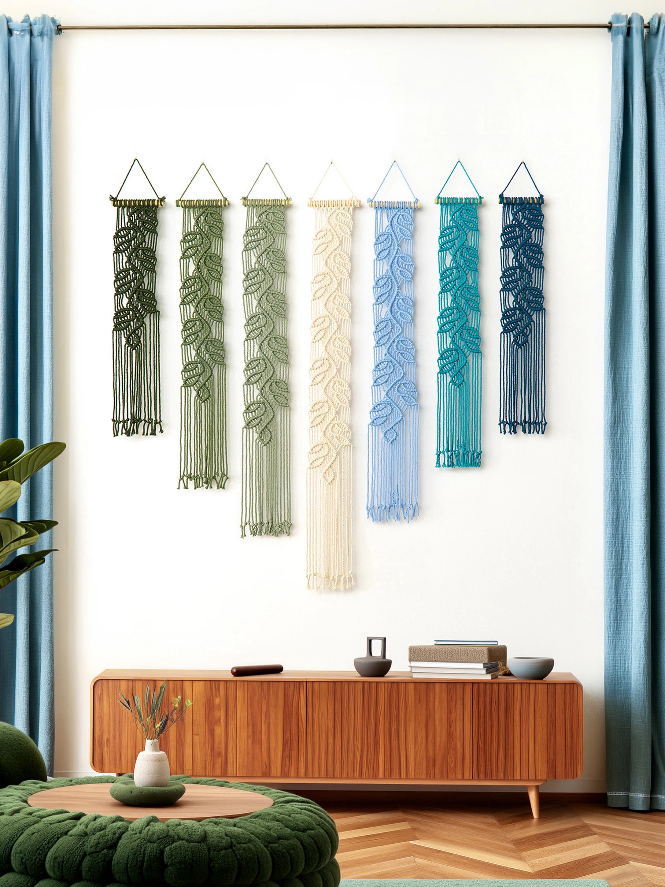 Six woven macrame wall hangings with leaf patterns in green, sage, cream, light blue, teal, and navy, displayed on a white wall above a wooden console table in a styled room.