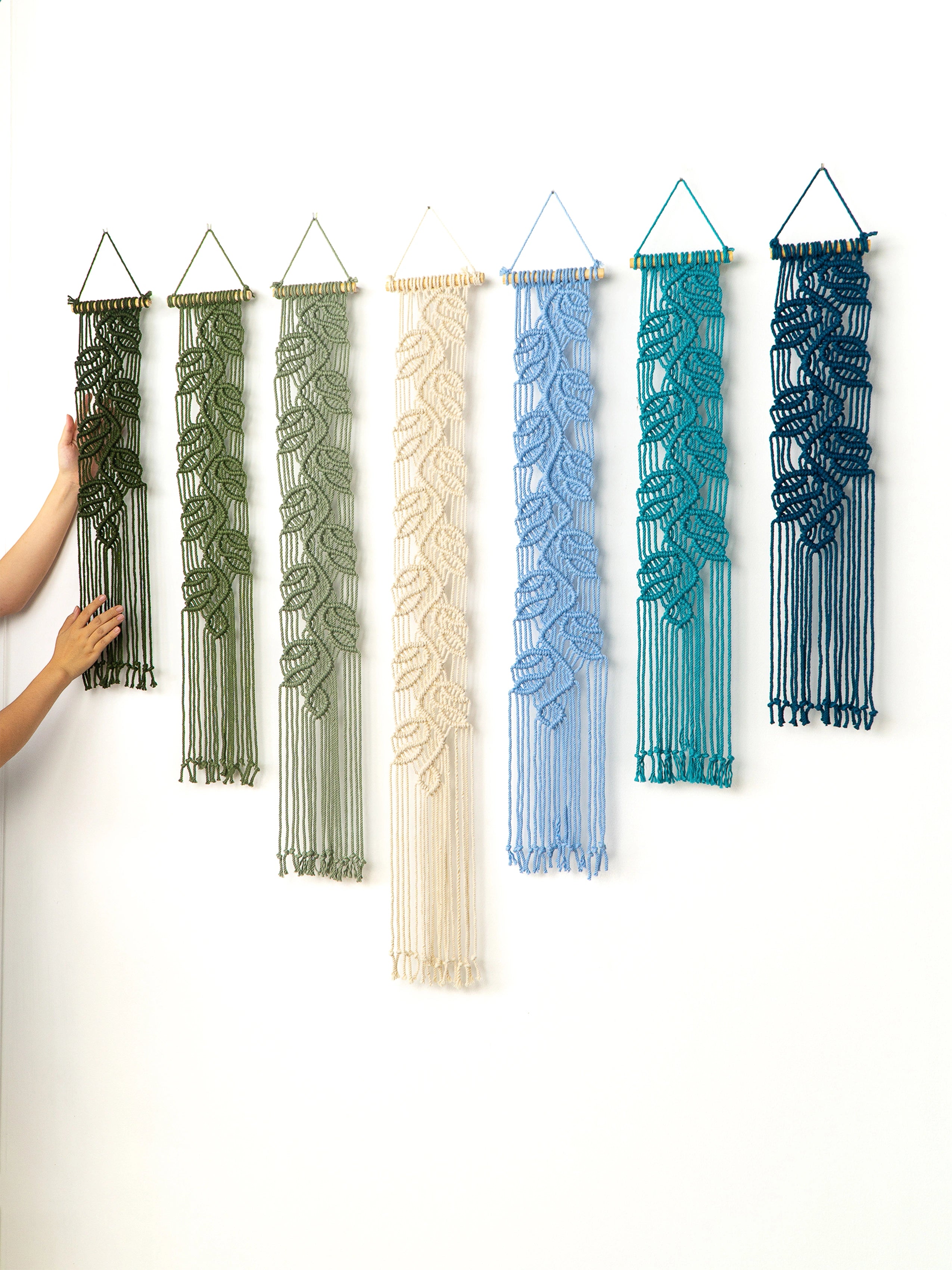 Six macrame wall hangings with leaf-like woven patterns are displayed on a white wall, shown in green, cream, light blue, teal, and navy. A person's hands are visible on the left, touching the first hanging.