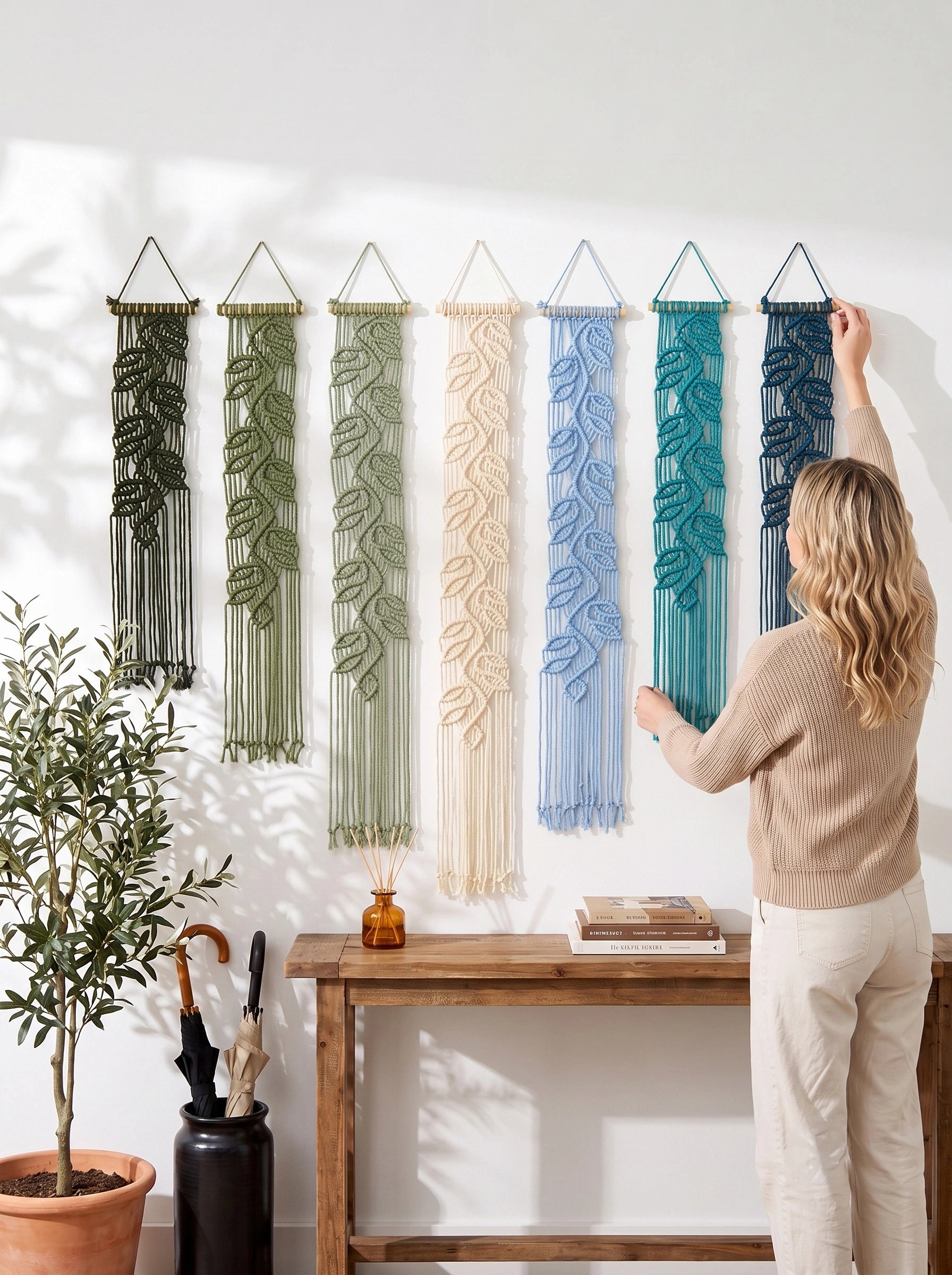 A woman hangs a blue leafy macrame wall hanging on a white wall next to five other leafy macrame wall hangings in green, olive, cream, light blue, and teal. A wooden console table and a potted plant are below.