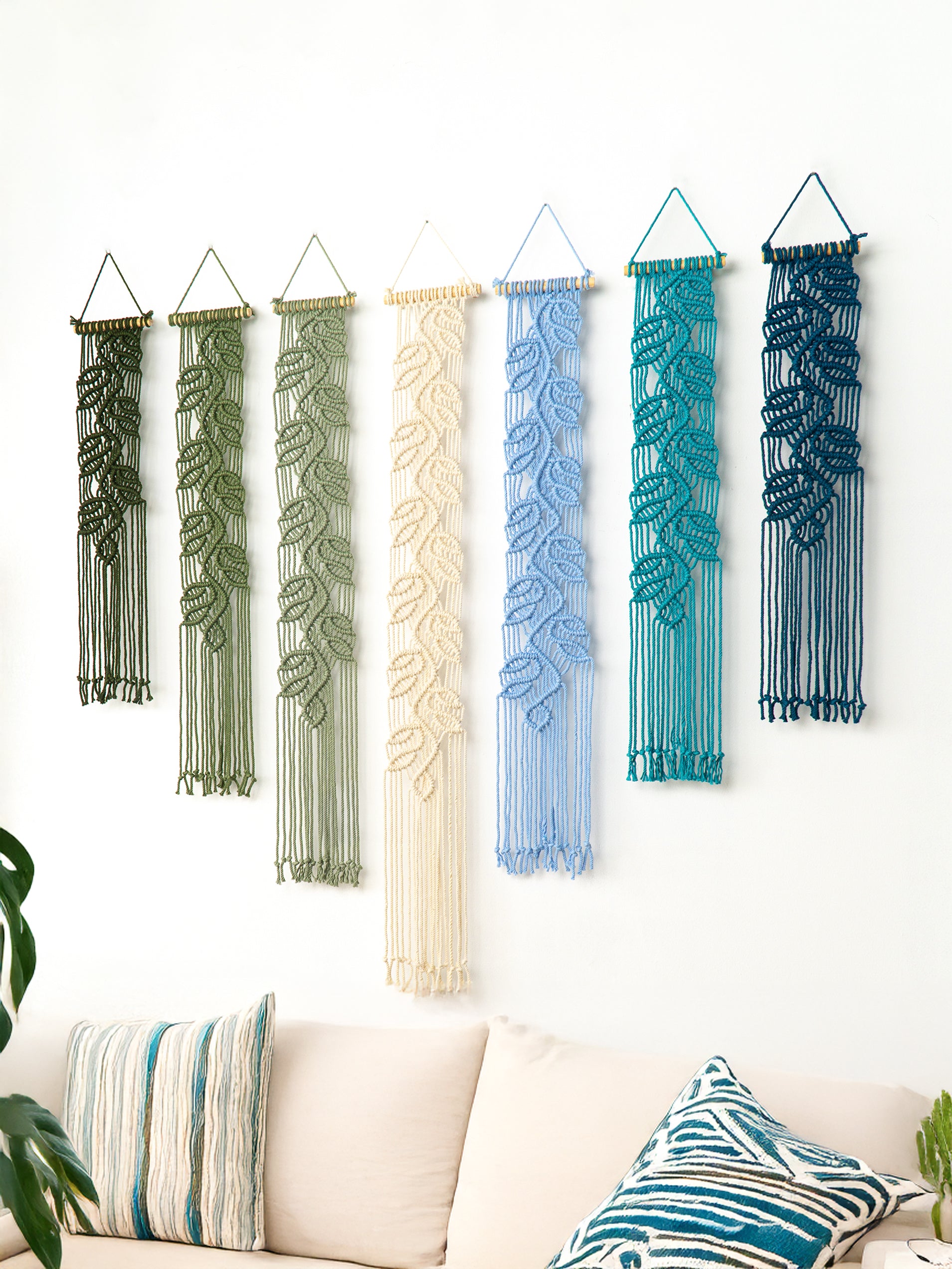 Set of six macrame wall hangings in various colors hanging on a white wall above a sofa.
