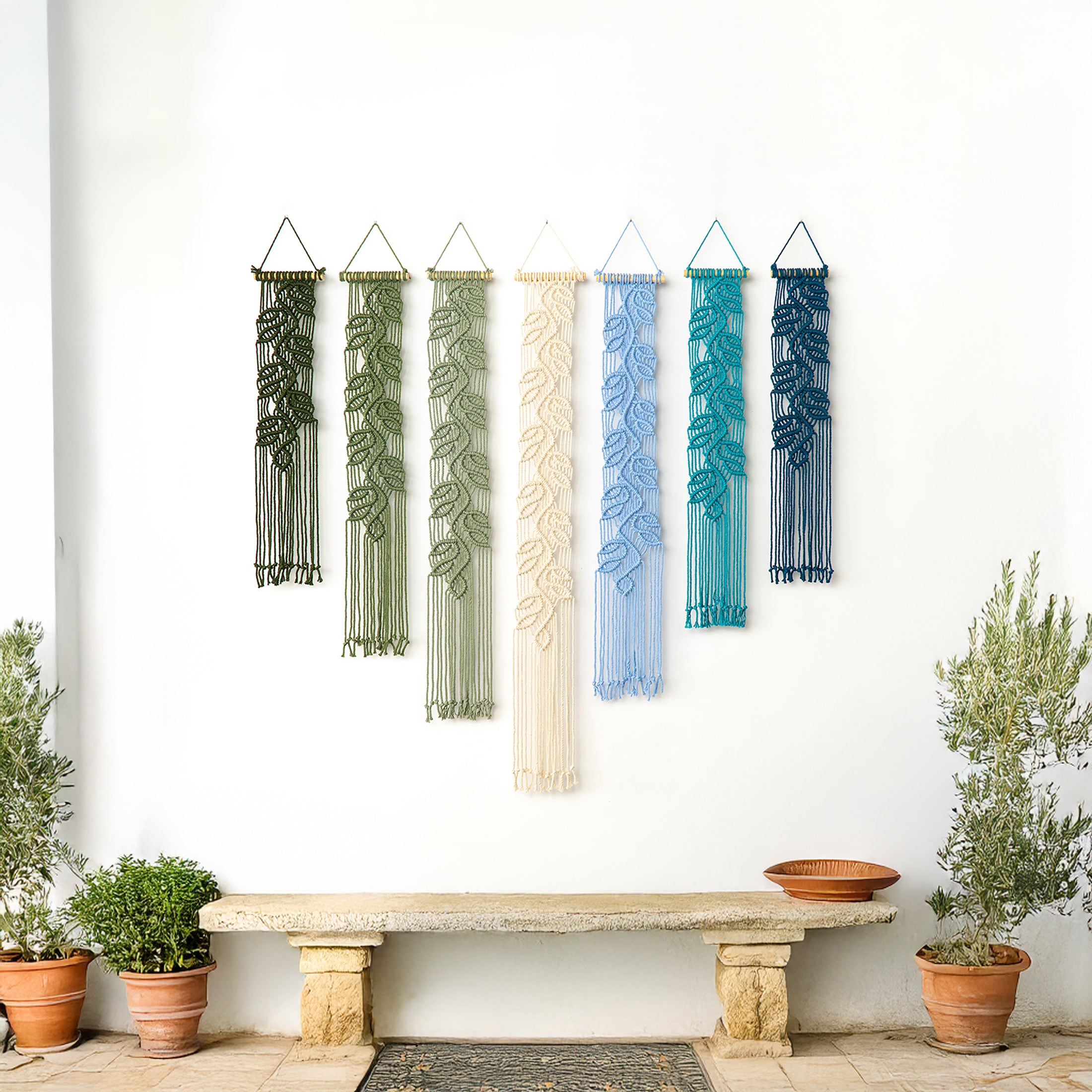 Five colorful wall hangings with leaf patterns hanging on a white wall above a stone bench.