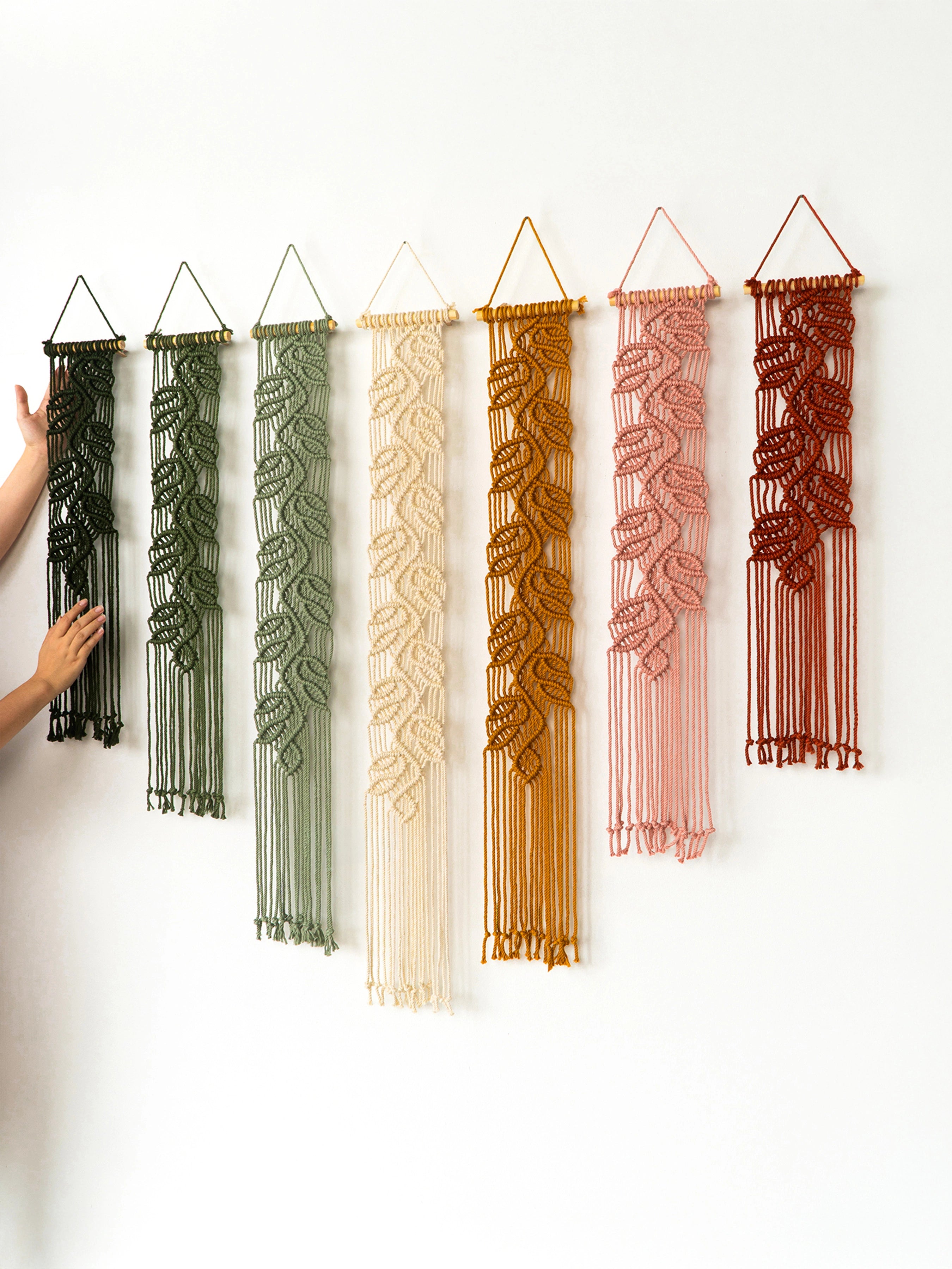 Six macrame wall hangings in different colors are displayed on a plain white wall, with a person's hand visible on the far left touching one of the hangings.