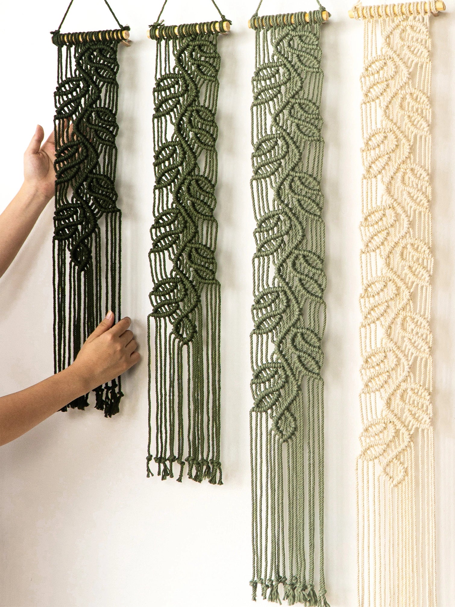 Three macrame wall hangings with leaf-like woven patterns hang on a white wall. Two hangings are dark olive green, and one is cream.