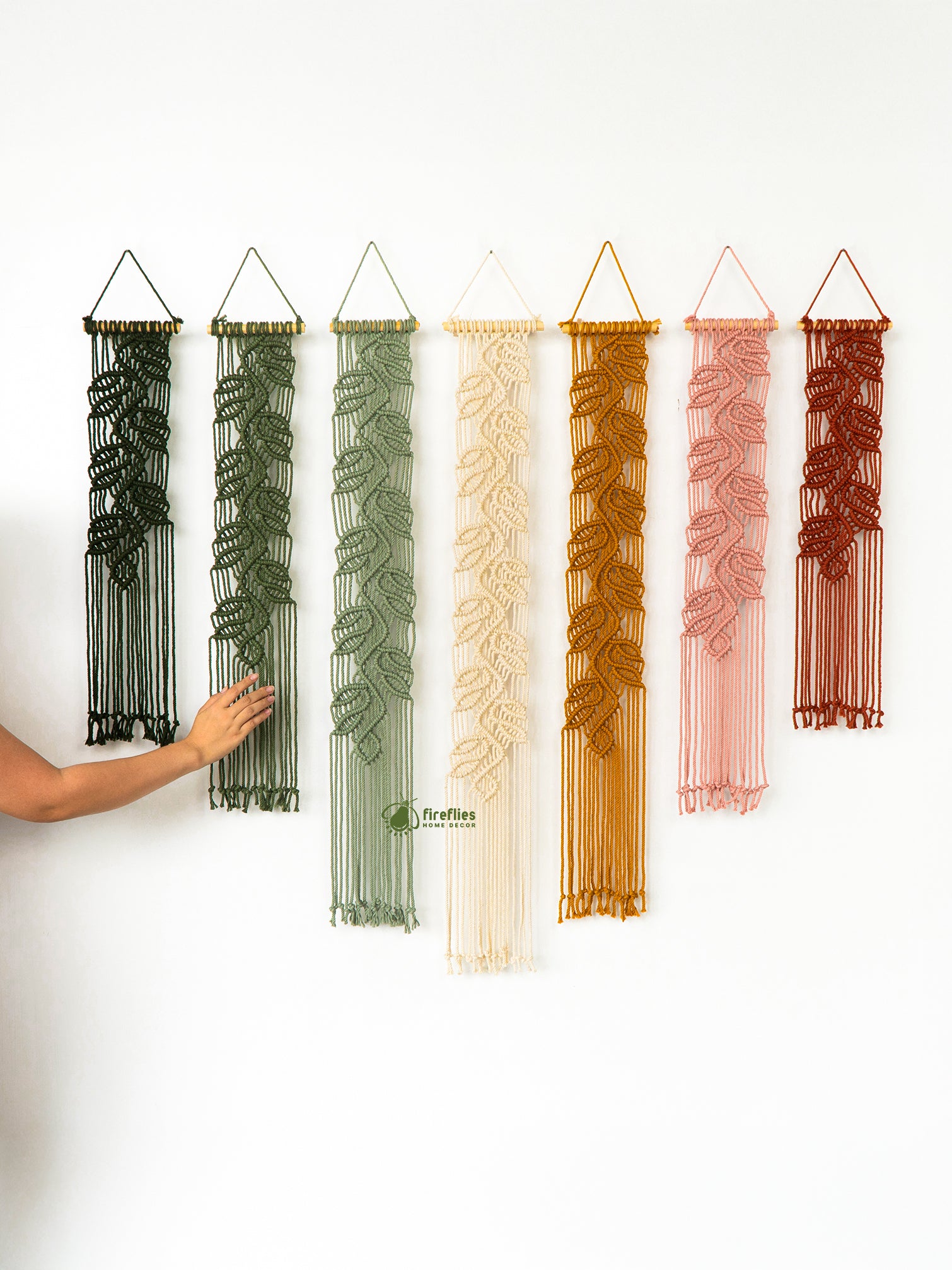 Set of six macrame wall hangings in various colors on a white wall.