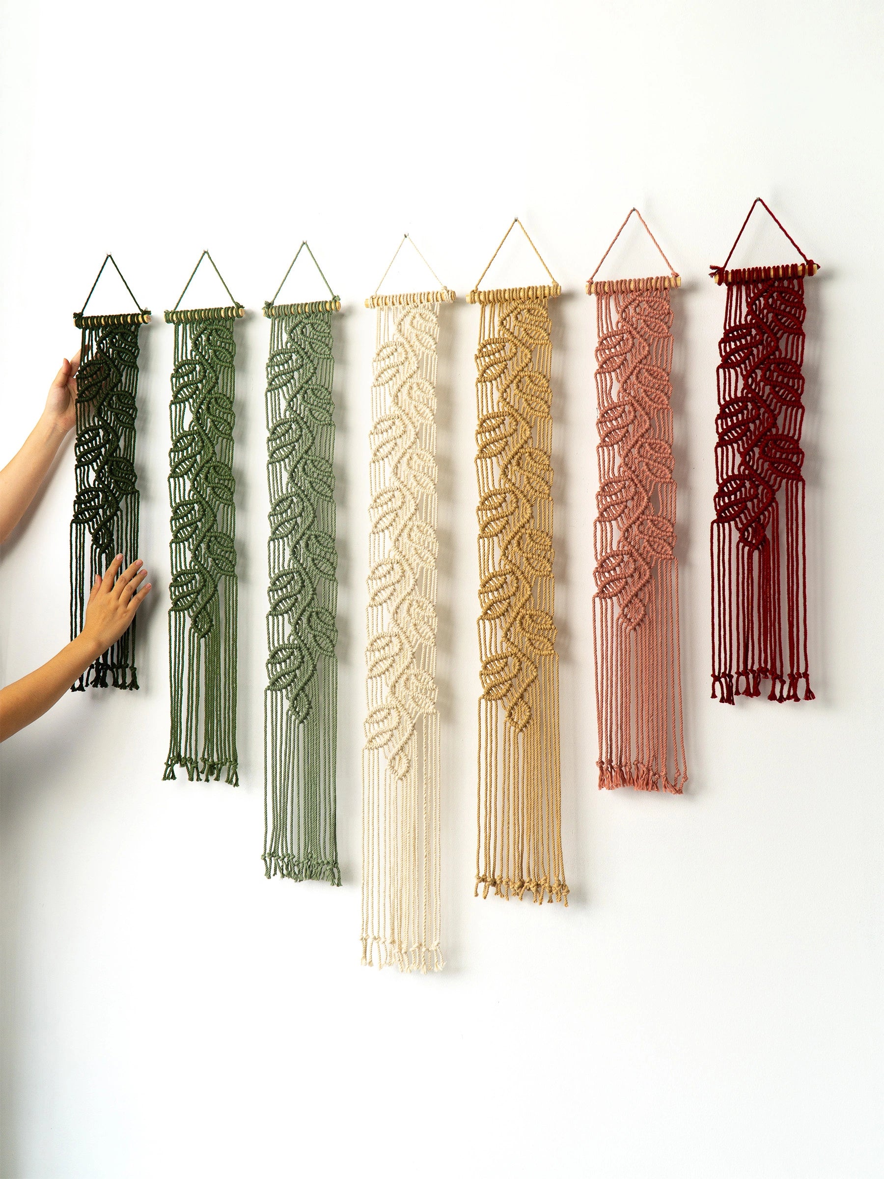 Six macrame wall hangings in different colors are displayed on a plain white wall. A person's arm and hand are visible on the left, touching the first hanging.