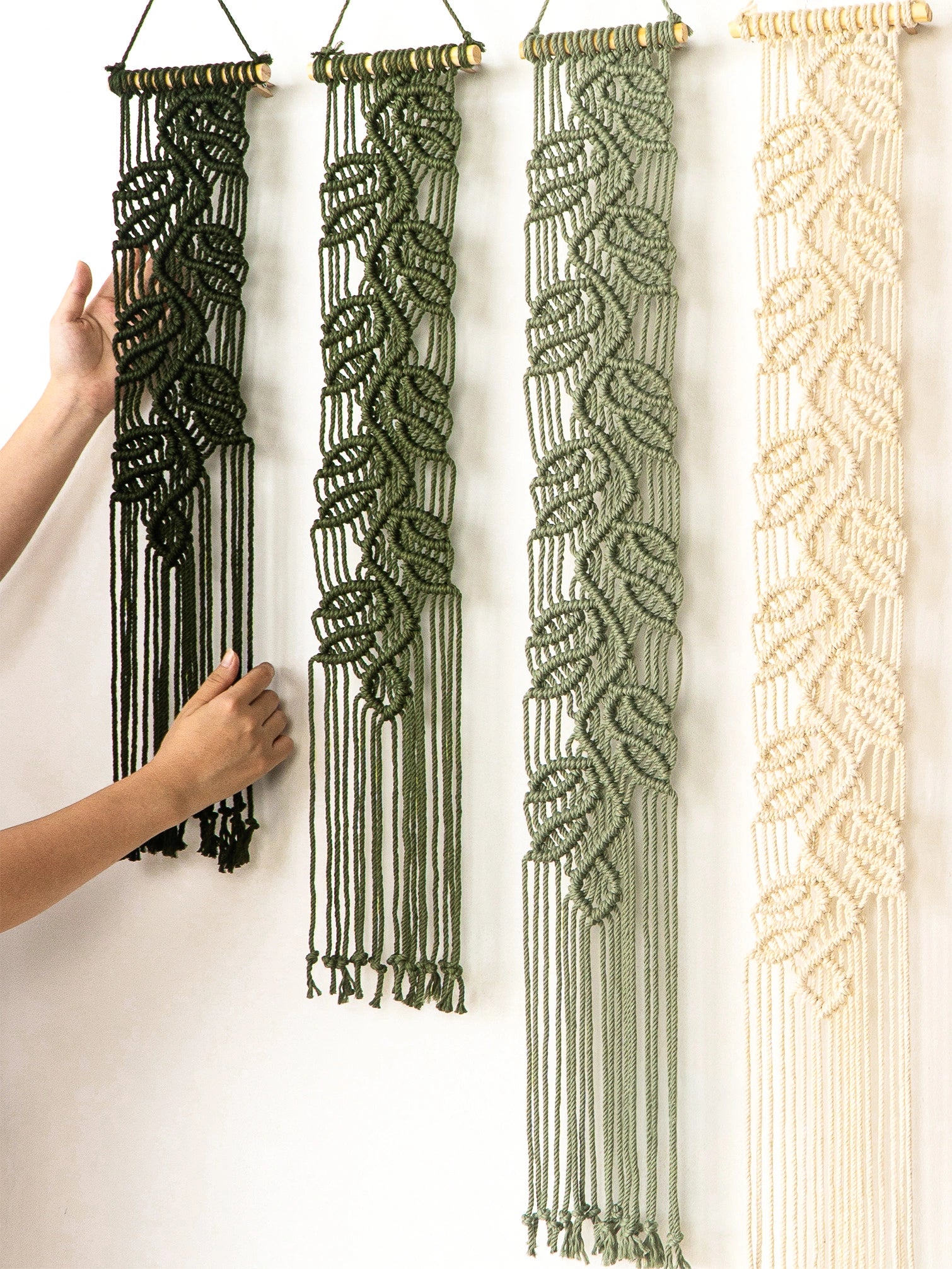 Four macrame wall hangings with leaf-like woven patterns are displayed on a white wall. Two hands are visible holding the leftmost dark green hanging to show its scale and texture.