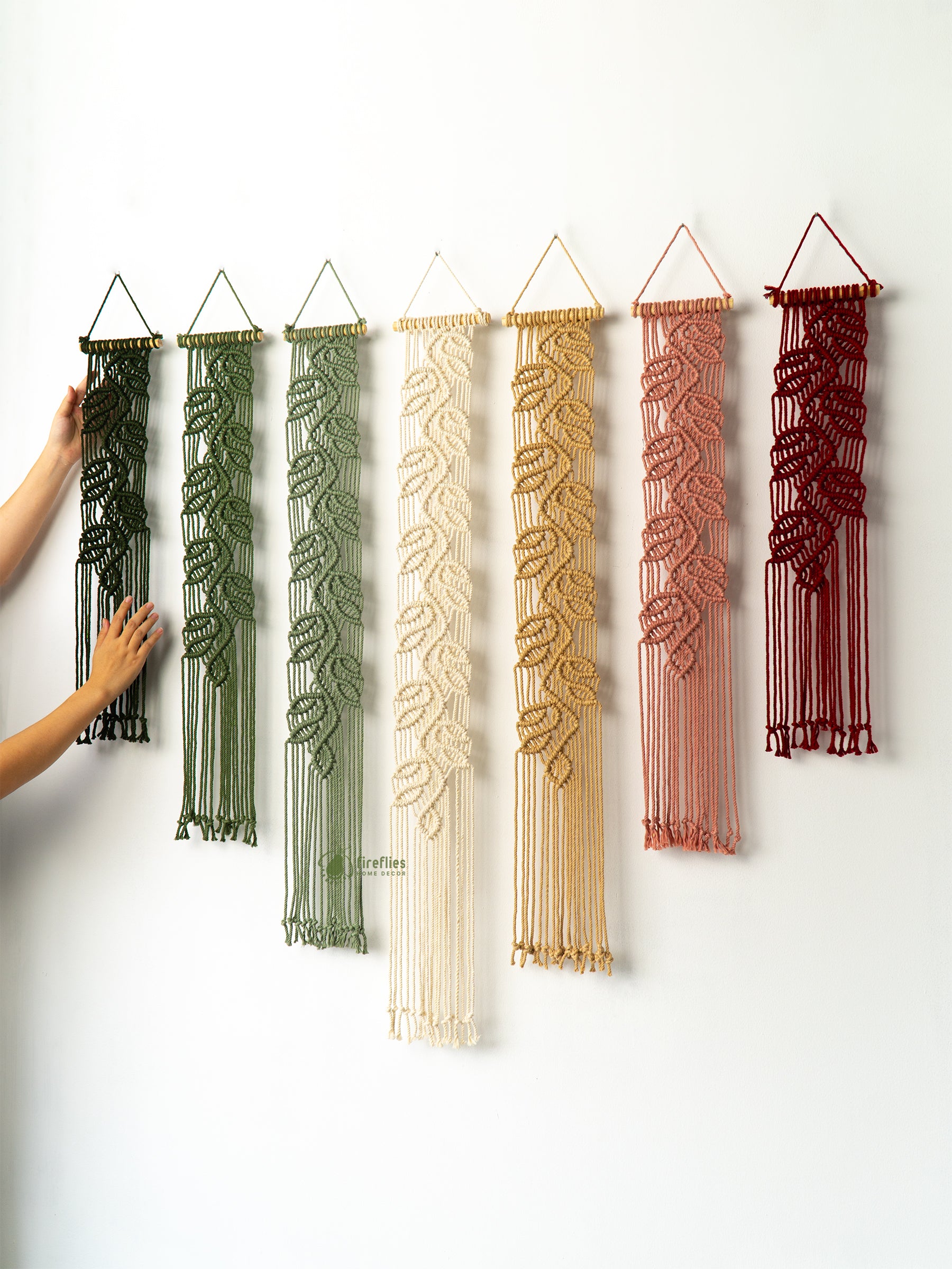 Set of six macrame wall hangings in various colors hanging on a white wall.