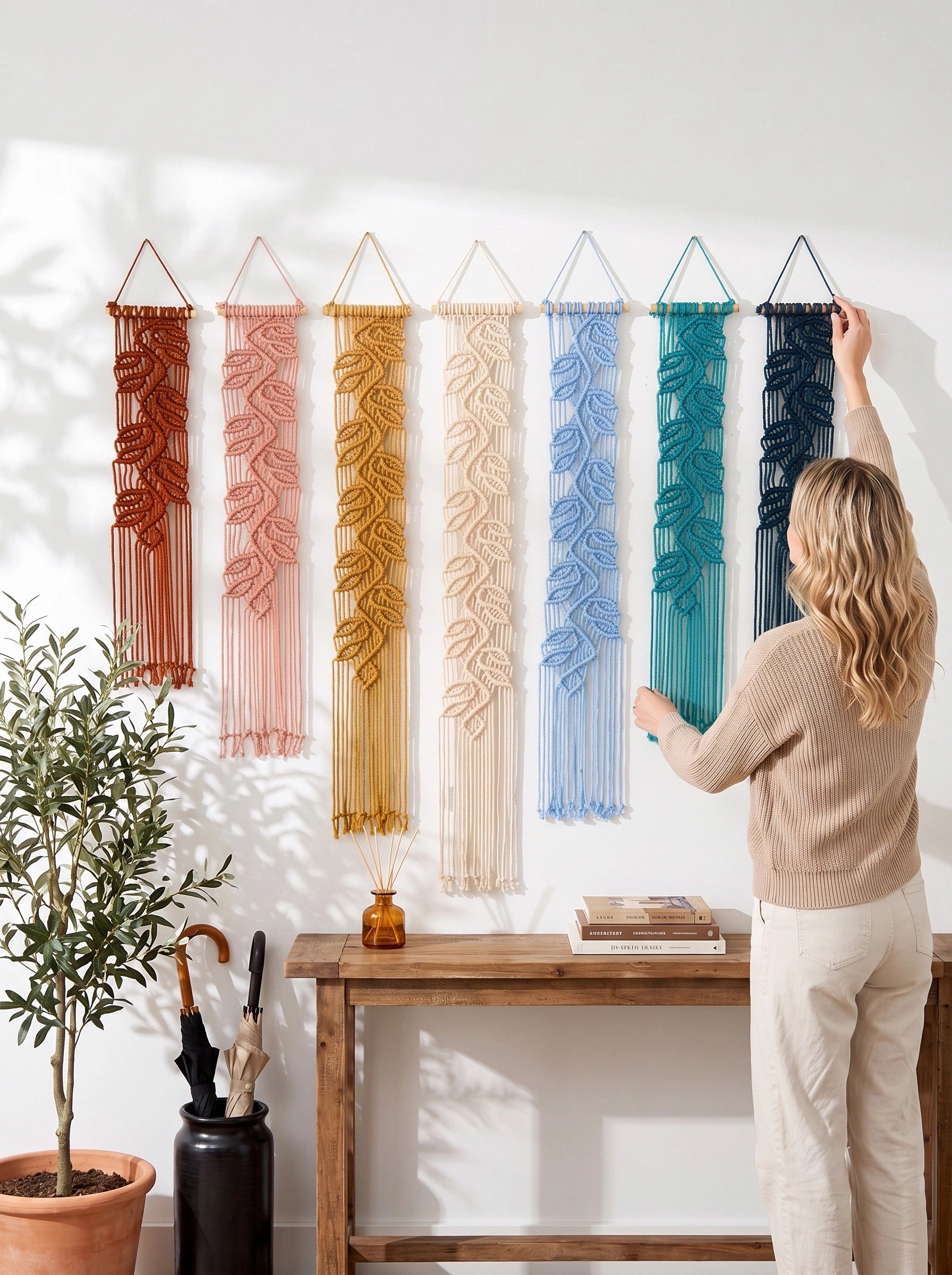 A woman hangs a dark blue macrame wall hanging on a white wall next to six other colorful woven wall hangings in terracotta, pink, mustard, cream, light blue, teal, and navy. A wooden console table with books and a potted plant sits below.