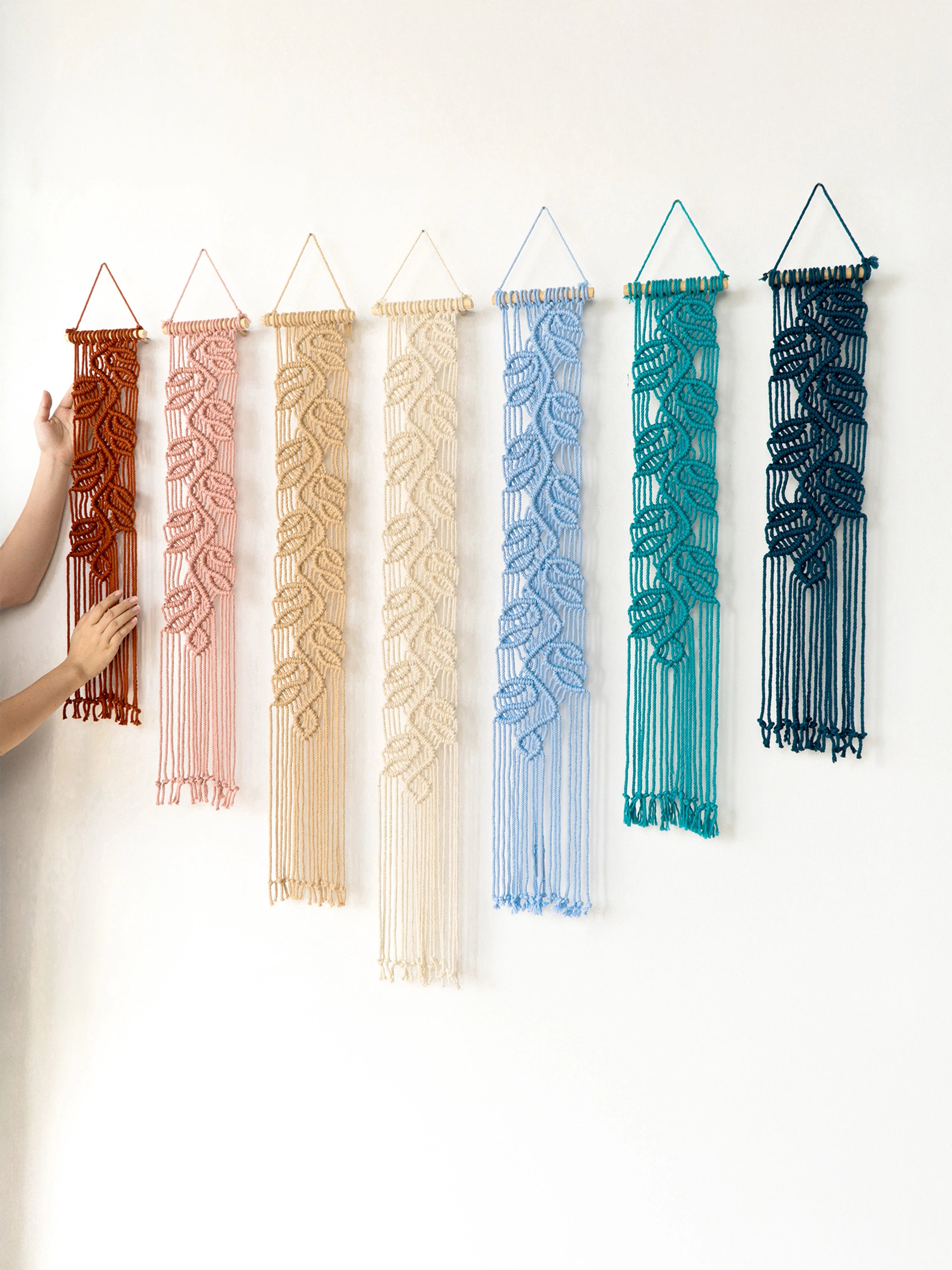 Six woven macrame wall hangings in different colors are displayed on a plain white wall. A person's hand is visible holding the leftmost orange-brown hanging, and the others are shown without hands.