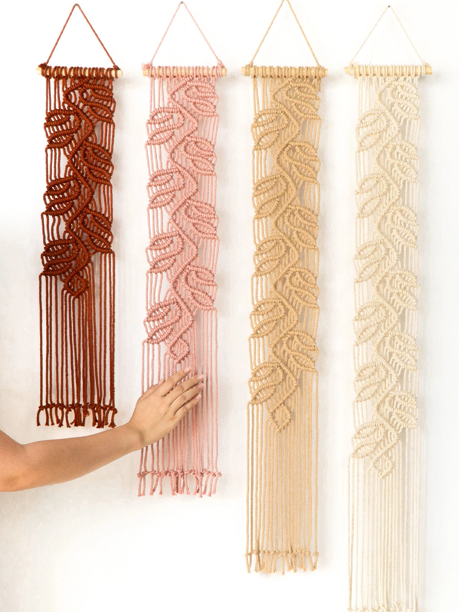 Four macrame wall hangings in rust, blush pink, tan, and cream are displayed against a white wall, with a hand reaching toward the pink one.