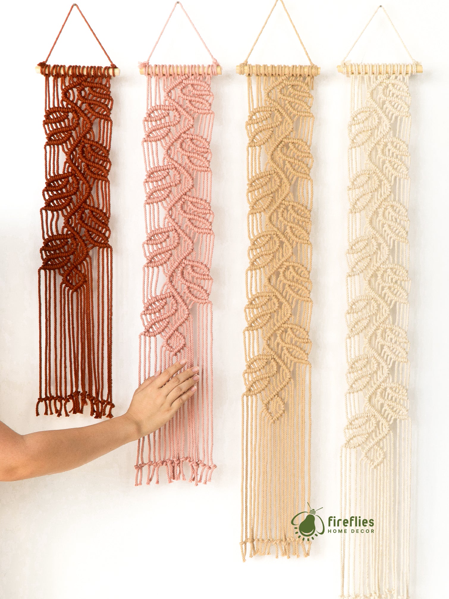 Four macrame wall hangings in different colors hanging on a white wall with a hand touching one of them.
