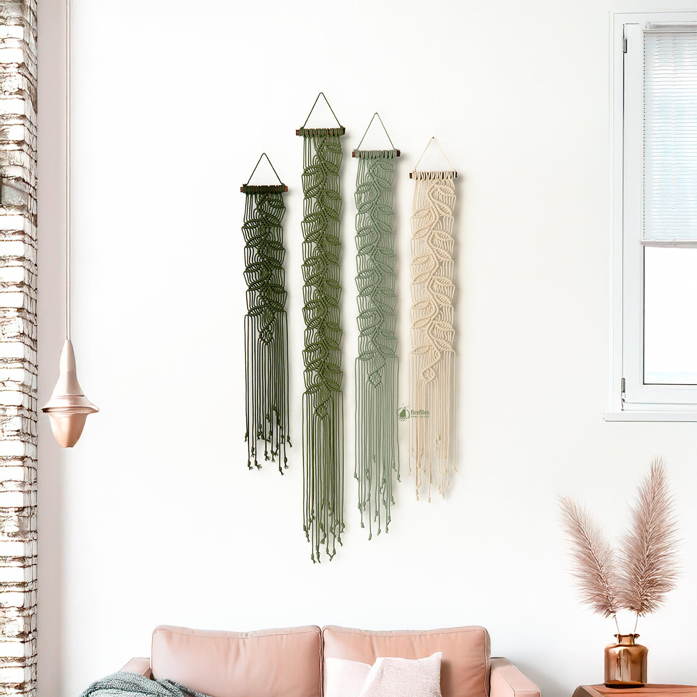 Three macrame wall hangings in green, beige, and brown hanging on a white wall above a pink sofa.